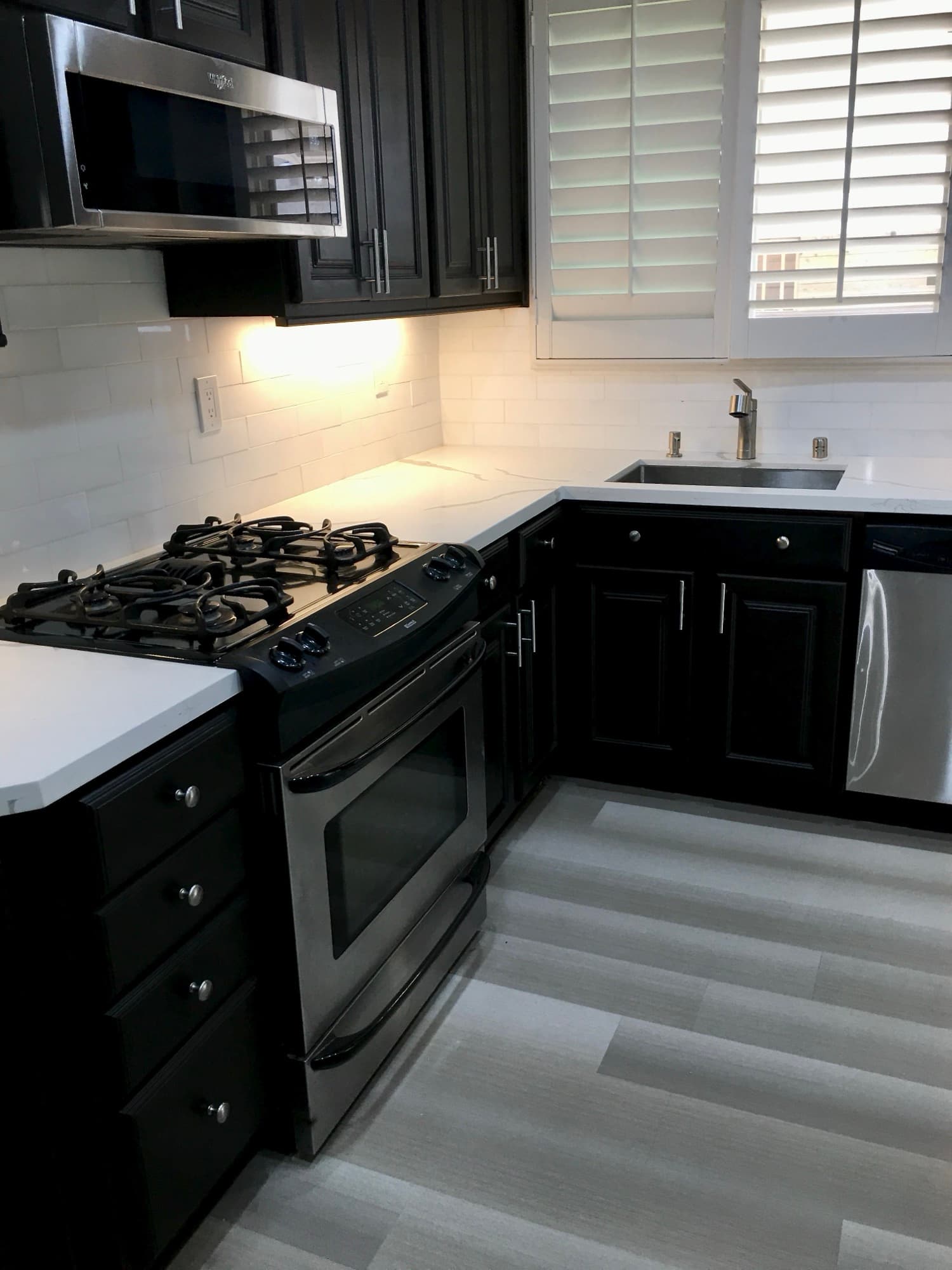 The same kitchen, black-cabinet finish, white quartz counter, subway tile, plantation shutters.